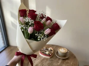 Punainen ruusukimppu ja vaaleat tulppaanit, kermapaperissa, kädessä kahvilapöydän yllä cappuccinon taustalla